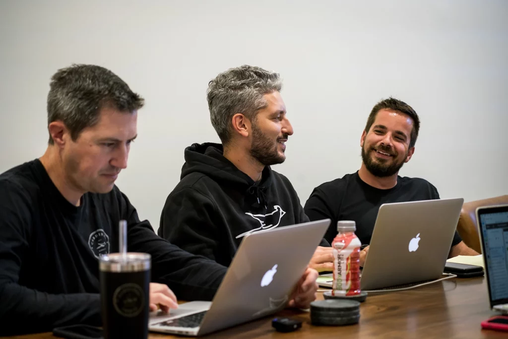 Three men sitting at a table looking at laptops and chatting